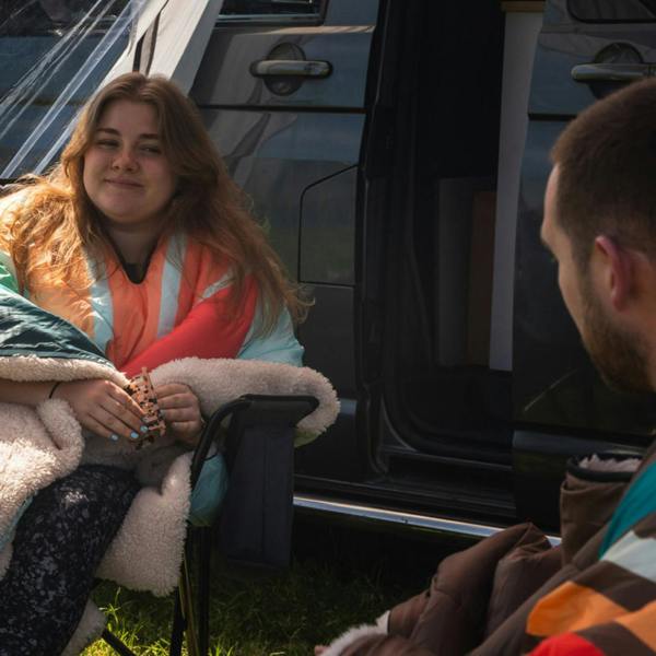 Insulated Outdoor Blanko OLPRO worn by a young lady and a young man, wrapped in the Blanko blankets whilst sat chatting, one wearing the insulated tonal tree tops with sherpa fleece and one wearing the vivid spruce. imageset:Tonal Tree Tops