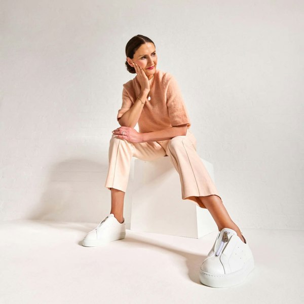 Woman sitting on a white stool wearing a pink outfit with white estella sneakers against a plain background