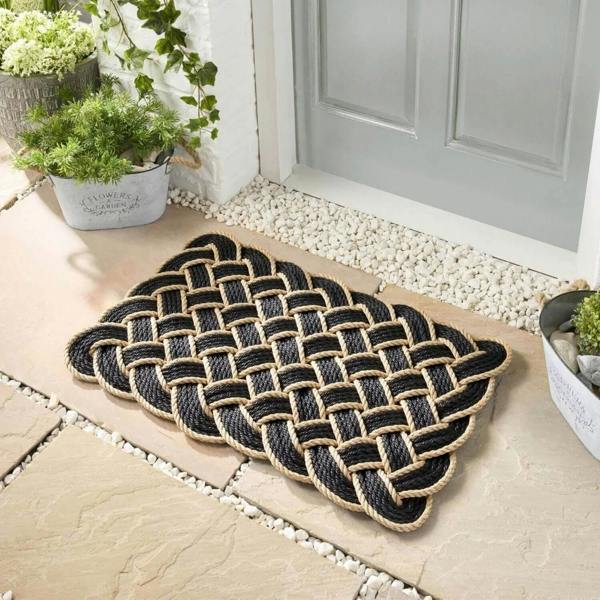 Woven doormat on a stone patio with plants and a door in the background