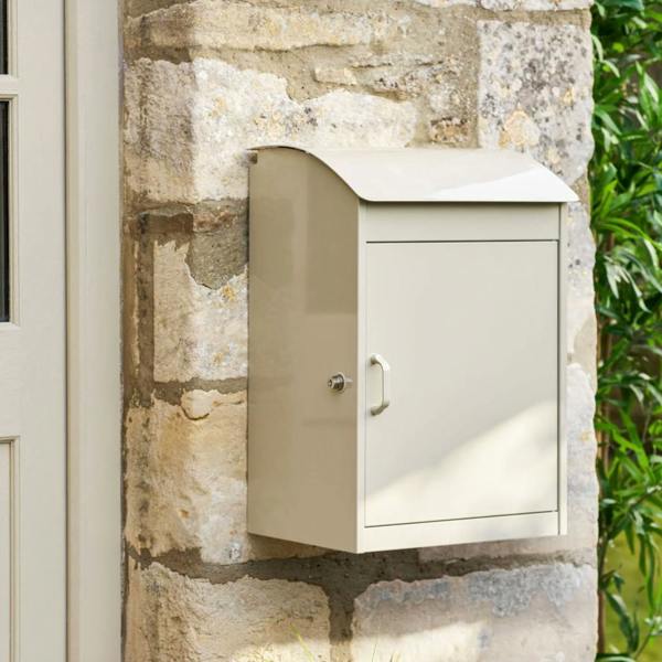 A neutral clay-coloured metal parcel box mounted on a rustic stone wall next to a white door.