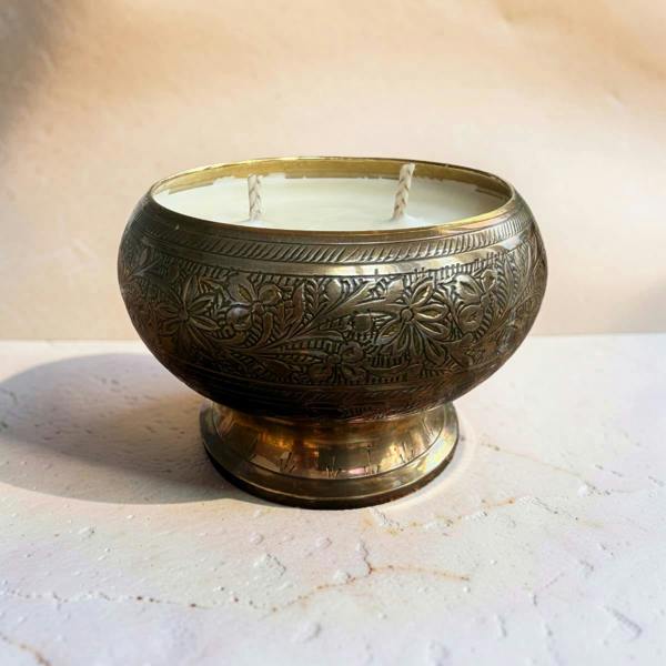 A double-wick soy candle in an ornate, hand-hammered brass bowl with floral engravings, sitting on a textured light stone surface.
