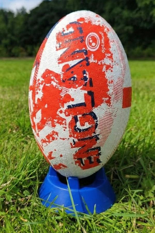 White and red Optimum England rugby ball with ENGLAND print upright on a blue kicking tee in grass - Optimum