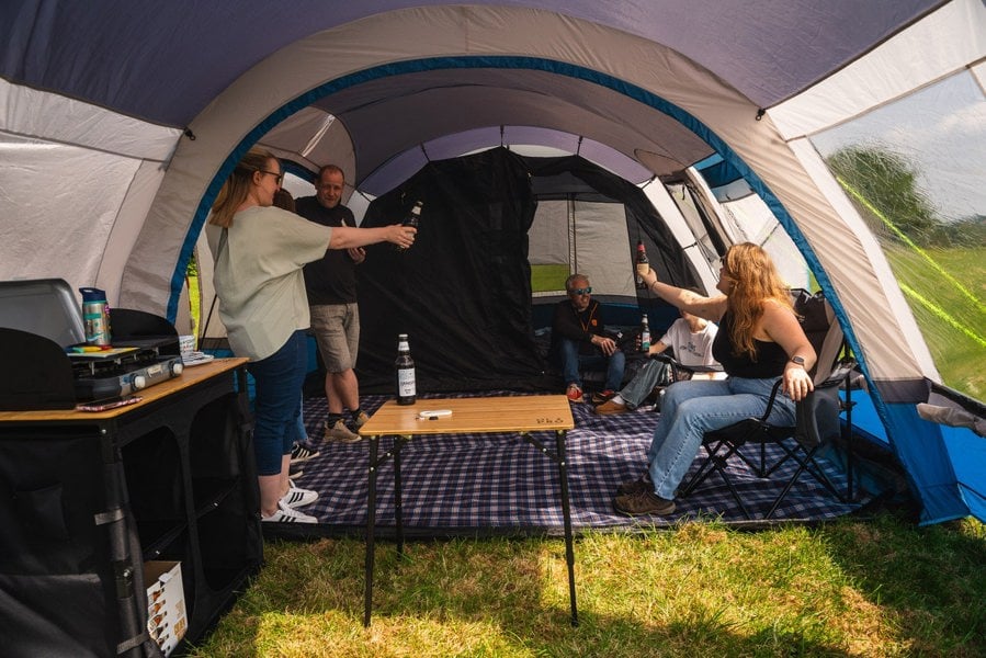 A group of friends sit inside the Poled Cocoon V2 Campervan Awning by OLPRO, some sat in camping chairs surrounding a bamboo folding table