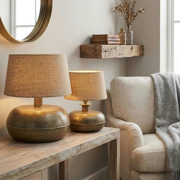 Small and large antique brass hammered table lamp bases styled on a rustic console table in a neutral cosy living room.