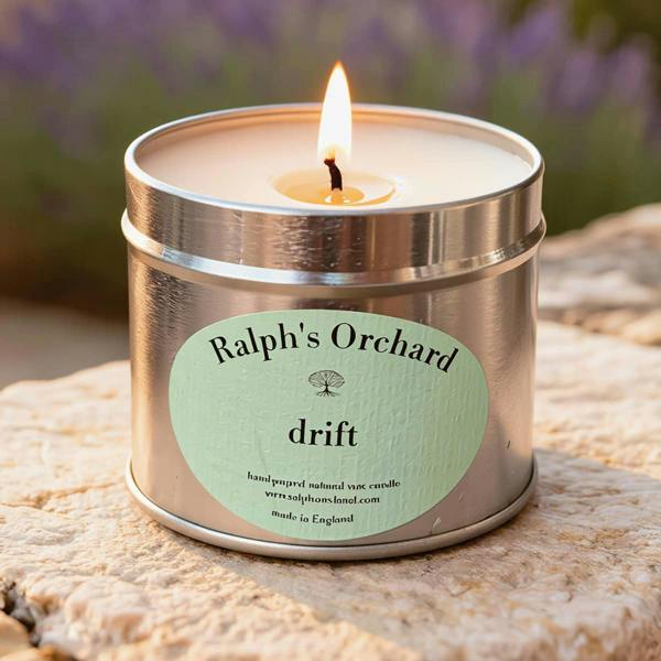 Candle in a metal tin labeled 'Ralph's Orchard drift' on a stone surface with blurred lavender in the background.