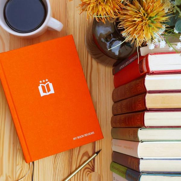 A vibrant orange cloth-bound My Book Reviews Journal on a wooden table next to a stack of books and a mug of coffee.