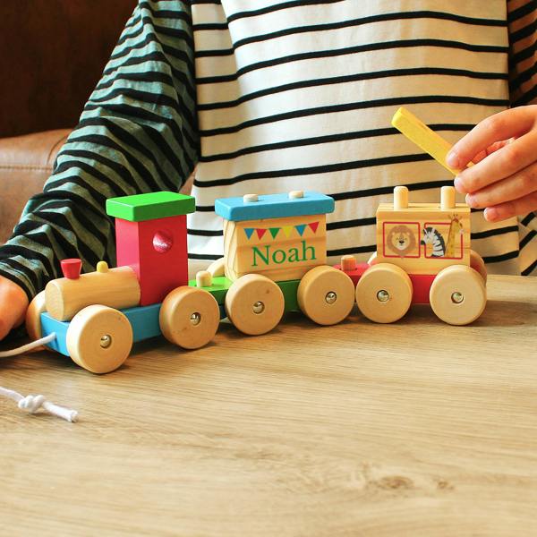 A wooden toy train with a circus theme sitting on a wooden table. A child's hand is holding a block above the train, which is personalised with the name "Noah".