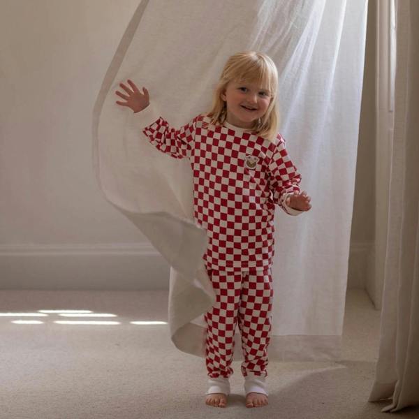 A smiling young toddler standing barefoot next to a white linen curtain, wearing a matching red and white checkerboard organic cotton Christmas pyjama set.