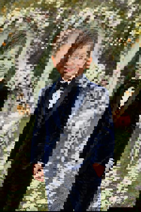 Young boy in a navy velvet suit standing in front of floral decorations at an outdoor event.