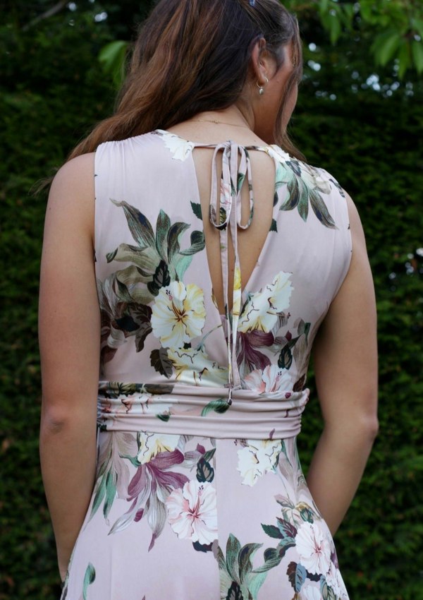 This is a young lady wearing a floral printed dress in pale pink. It has a mock V neck style top, which has gathers under the bust and round the back and the shoulders with an open back with a tie, secured into a bow. The model is facing away from us and looking over her shoulder, with her hair swept away to show the detail in the back. She is silhouetted against a leafy garden background.