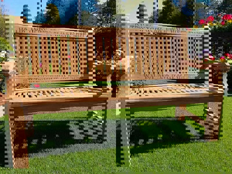 A wooden memorial bench with engraved text on the top rail, placed on a grassy area with flowers in the background.