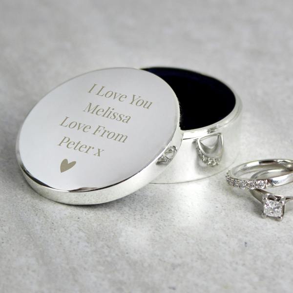 A round silver plated ring box sits open on a stone surface next to two silver diamond rings. The lid is laser-engraved with the romantic custom message 'I Love You, Melissa, Love From Peter x' positioned above a small heart icon.