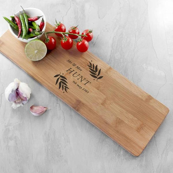 A long rectangular bamboo serving board engraved with Mr and Mrs HUNT 9th May 1985 and leafy branches, styled on a stone surface with fresh vine tomatoes, garlic, and a bowl of green chillies.