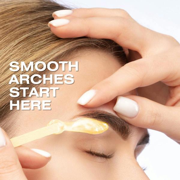 A close-up of a person’s closed eye as another applies Rio Honey Go Smooth Stripless Hard Wax Beads (100g) with a wooden stick to shape their eyebrow. White text reads, “SMOOTH ARCHES START HERE.”.