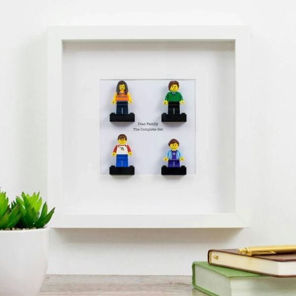 A white picture frame hanging on a wall, featuring four personalised mini figures (a woman, a man, a boy, and a girl) standing on black brick bases, with the text "Diaz Family" and "The Complete Set" printed on the white backing.