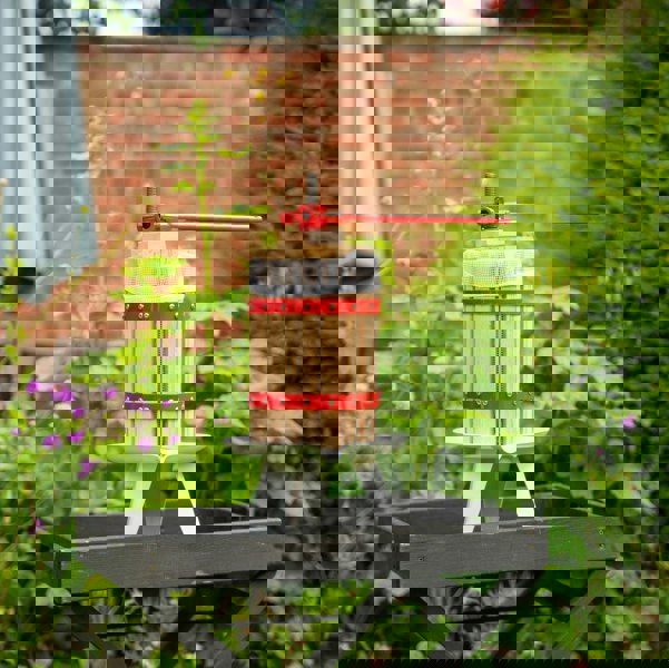 KuKoo 6 Litre Fruit Press with wooden barrel, red metal accents, and a sturdy base, set against a lush garden backdrop
