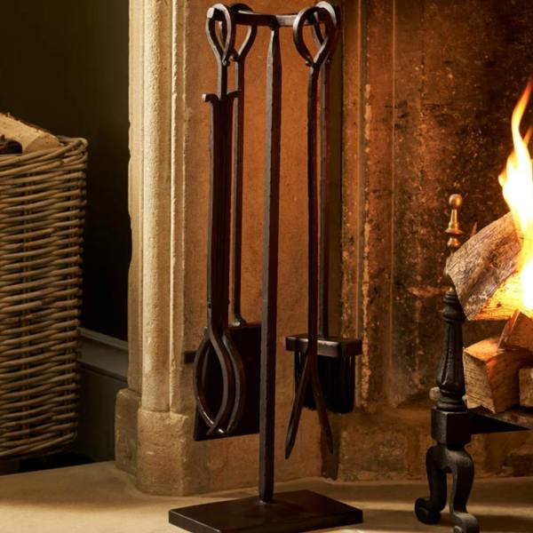 A wrought iron fireside companion tool set with an antiqued brown finish standing next to a stone fireplace with a roaring log fire.
