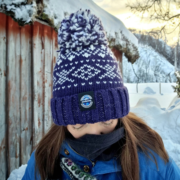  Woman at the side of a log cabin in Norway is wearing a chunky Nordic knit bobble hat. The navy and white Nordic design hat also has a deep ribbed headband, contains reflective yarns and has a luxury sherpa fleece lining.