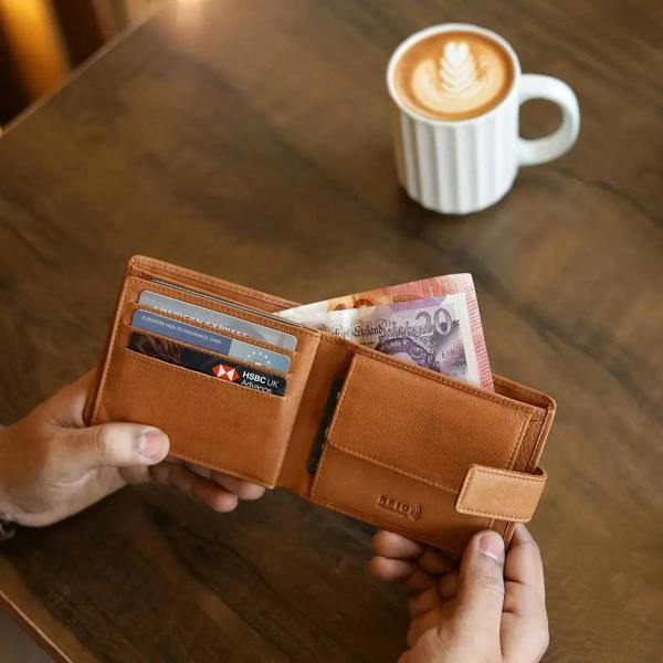 Cognac leather wallet with money and cards held by hands on a wooden table with a coffee cup in the background.