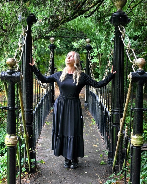 The young woman in the picture has long blonde hair and she is standing on a bridge in a park with lots of trees and foliage. She is holding onto the poles at the entrance to the bridge and is glancing upwards to her right and is facing the camera. She is wearing a boho gathered midi skirt with pockets in black. She is also wearing our beatnik top with crew neck in black