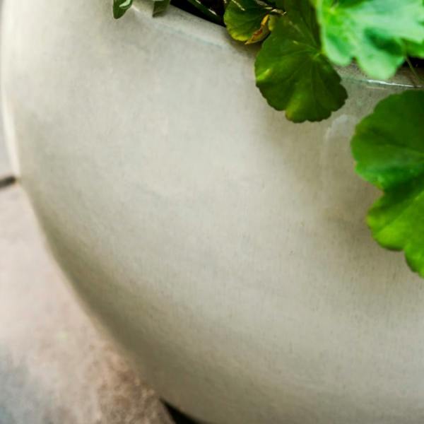 Close-up macro detail of the smooth, glossy white speckled glaze on a round terracotta garden planter with green foliage draped over the edge.