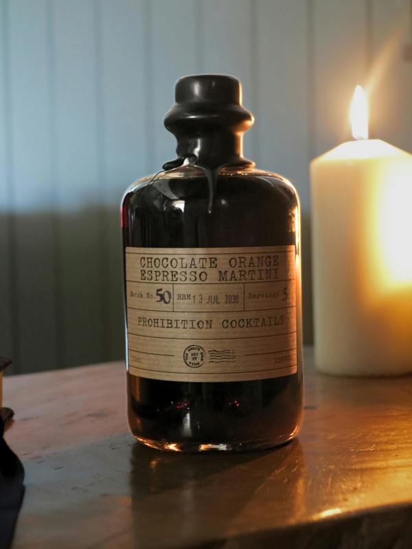 A dark glass bottle labeled 'Chocolate Orange Espresso Martini' by Prohibition Cocktails sits on a wooden table next to a lit candle in a warm, dimly lit setting.