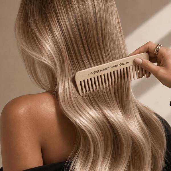 A close-up lifestyle shot of a person with long, wavy blonde hair using a light beige wooden comb branded '# ROSEMARY HAIR OIL co.' The hand holding the comb is visible against a warm beige background.