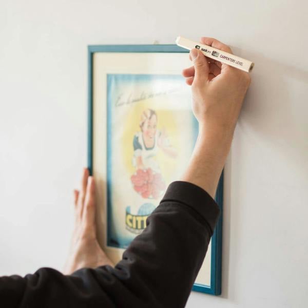 A close-up of a person using a cream-coloured carpenter's spirit level pencil to align a picture frame on a white wall.