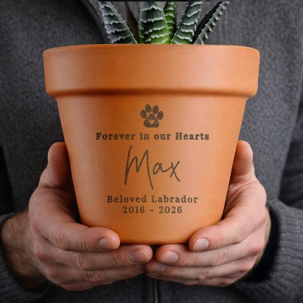 Hands holding a personalised terracotta pet memorial plant pot engraved with a pawprint, Forever in our Hearts, Max, Beloved Labrador, 2016 - 2026.