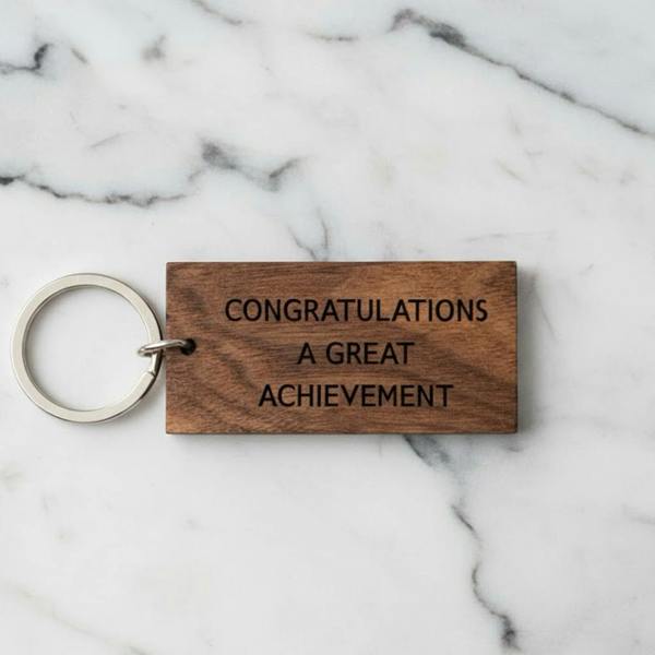A rectangular solid walnut wood keyring engraved with the text "CONGRATULATIONS A GREAT ACHIEVEMENT" resting on a white marble surface.