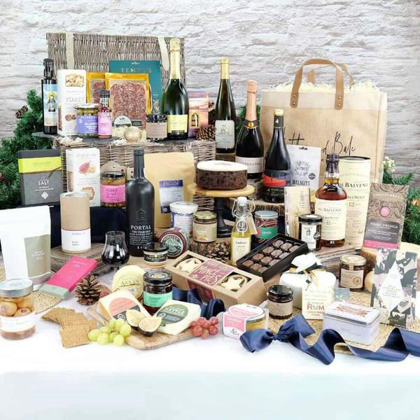 Grand festive display of luxury Christmas food hamper products including champagne, port, cheeses, charcuterie, and cakes set around a wicker basket and Hessian bag