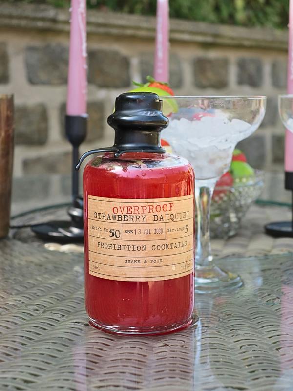 A clear glass bottle filled with bright red liquid, featuring a vintage-style tan label that reads 'Overproof Strawberry Daiquiri' and 'Prohibition Cocktails,' set on a metal patio table with a cocktail glass and pink candles in the background.