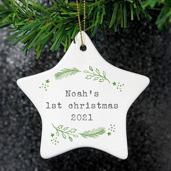 A white personalised star-shaped ceramic Christmas decoration hanging from a tree branch with the custom text Noah's 1st christmas 2021 surrounded by green holly and pine graphics.