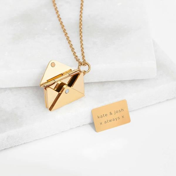 A classic gold envelope pendant necklace from Abode Home & Gifts open on a marble tier next to an engraved message insert reading 'kate & josh x always x'.