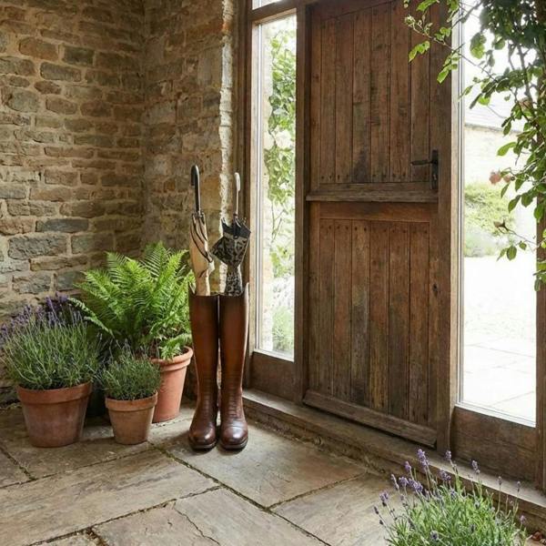 A leather-effect riding boot umbrella stand holding two brollies, styled on a rustic flagstone floor in a stone-walled cottage porch next to potted lavender and fern plants by an open wooden door.