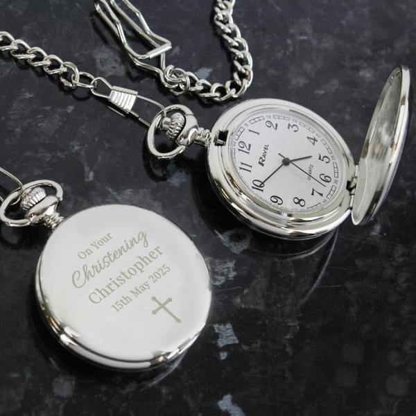 A silver pocket watch with a chain lying on a dark surface. The watch is engraved with the text "On Your Christening," "Christopher," "15th May 2025," and a cross symbol. A second watch is shown open next to it, revealing a white clock face.