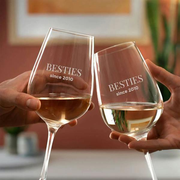 Two hands clinking two custom engraved wine glasses from Abode Home & Gifts that read 'BESTIES since 2010' filled with white wine.