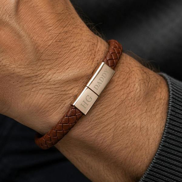 A close-up view of a man's wrist wearing the braided tan leather charger bracelet with the gold metallic clasp engraved with HG and DADDY.
