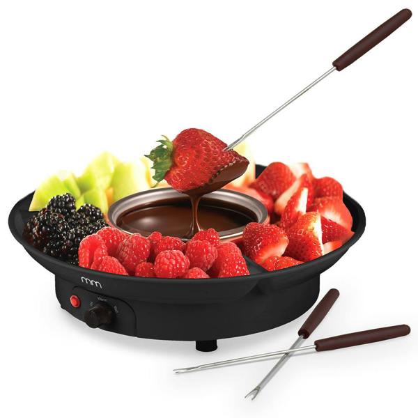 A studio product shot of a black electric fondue set against a clean white background. The central pot is filled with rich melted milk chocolate, with a fresh strawberry on a fondue fork being dipped into it. The segmented outer plastic tray compartments are generously piled with fresh fruit, including raspberries, sliced strawberries, blackberries, and honeydew melon chunks. Two extra dipping forks rest beside the active electrical base.