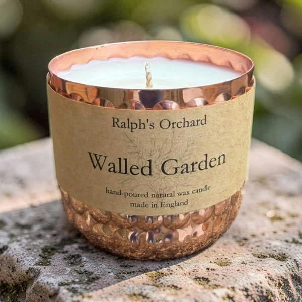 A Ralph's Orchard 'Walled Garden' natural wax candle in a hammered copper glass jar, resting on a textured stone surface with a blurred garden background.