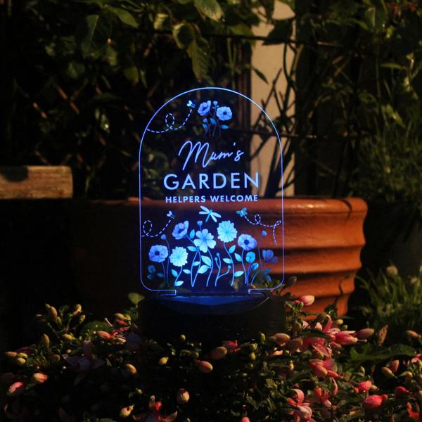 An arched clear acrylic garden solar stake glows with a bright blue LED light at night. The sign is precision-etched with a three-line custom text reading 'Mum's GARDEN, HELPERS WELCOME' framed by detailed wildflower graphics, bumblebees, and a dragonfly. The base is positioned in a flowering outdoor bush next to a rustic terracotta garden pot.