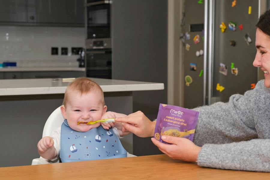 Baby being fed by an adult in a kitchen setting with a more toddler meal sweet potato dhal