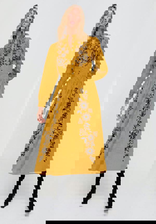 Woman wearing a mustard yellow dress with black floral embroidery on a white background