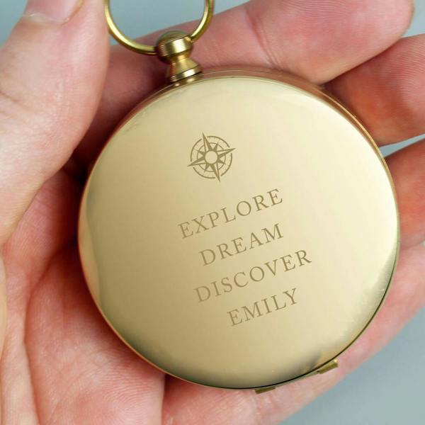 A close-up shot of a round, brushed-brass metal pocket compass being held in a hand, engraved with a small compass rose motif followed by the uppercase words 'EXPLORE DREAM DISCOVER EMILY'.