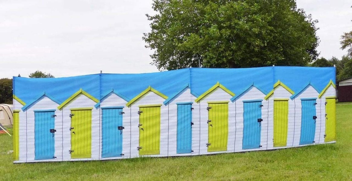 Beach Huts Design 4 Pole Compact Windbreak Steel Poles by OLPRO pictured on a field. It's Blue sky with White Beach huts with alternate blue and yellow doors fully assembled.