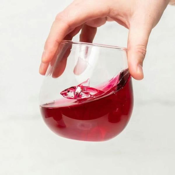 A hand swirling red liquid inside a clear glass tumbler to reveal a glass shark sculpture emerging from the beverage.