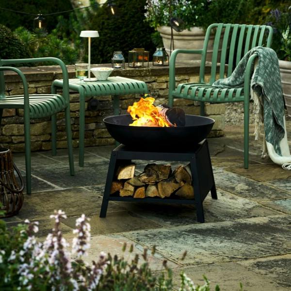Matte black steel fire bowl with a burning log, featuring an integrated base for log storage, styled on a stone patio between two green garden chairs.