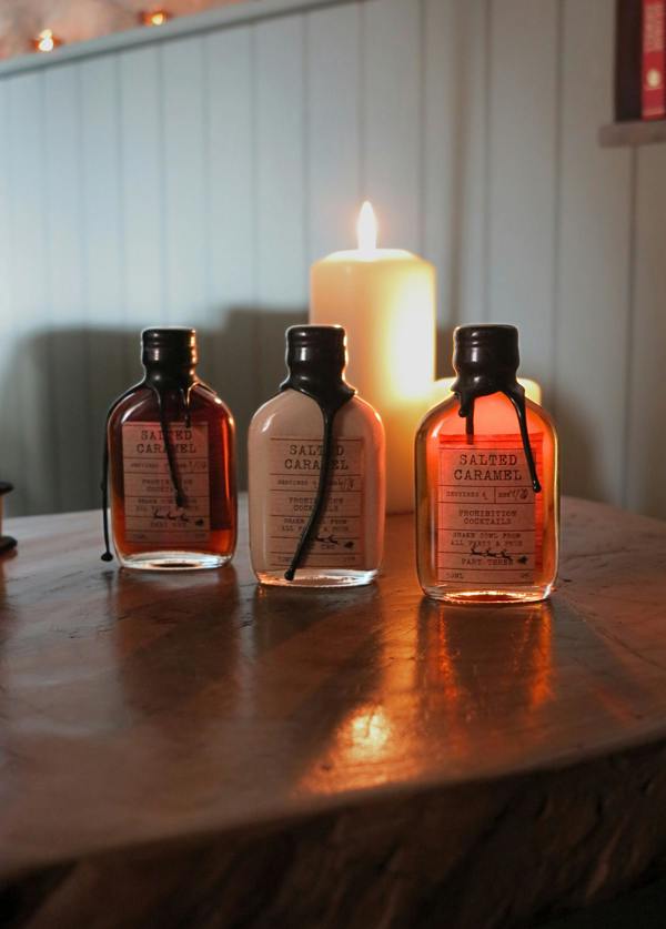 Three small amber glass bottles with black caps and cream labels reading 'SALTED CARAMEL' are arranged on a wooden surface in front of a glowing white pillar candle.