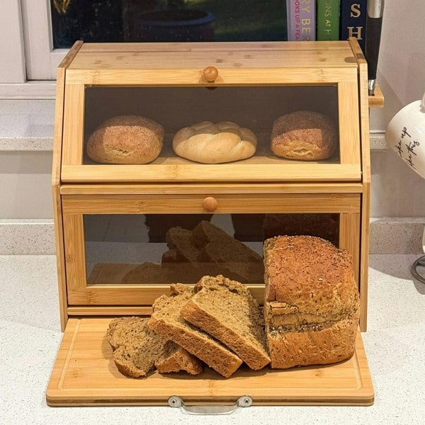 A wooden bamboo bread box with two transparent compartments showing various breads inside. The bottom is open with sliced whole grain bread and a seeded loaf displayed on the slide-out cutting board.