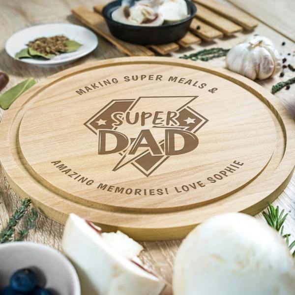 A round wooden chopping board engraved with a central "Super Dad" logo and a personalised message around the edge that reads "Making Super Meals & Amazing Memories! Love Sophie," surrounded by fresh cooking ingredients like garlic, mushrooms, and herbs.